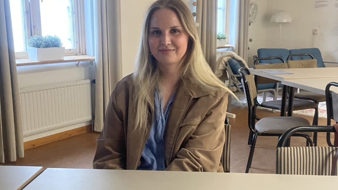 Det sitter en mörkblond tjej med en brun läderjacka med en blåskjorta under. Hon sitter i ett ljust klassrum och ler.
