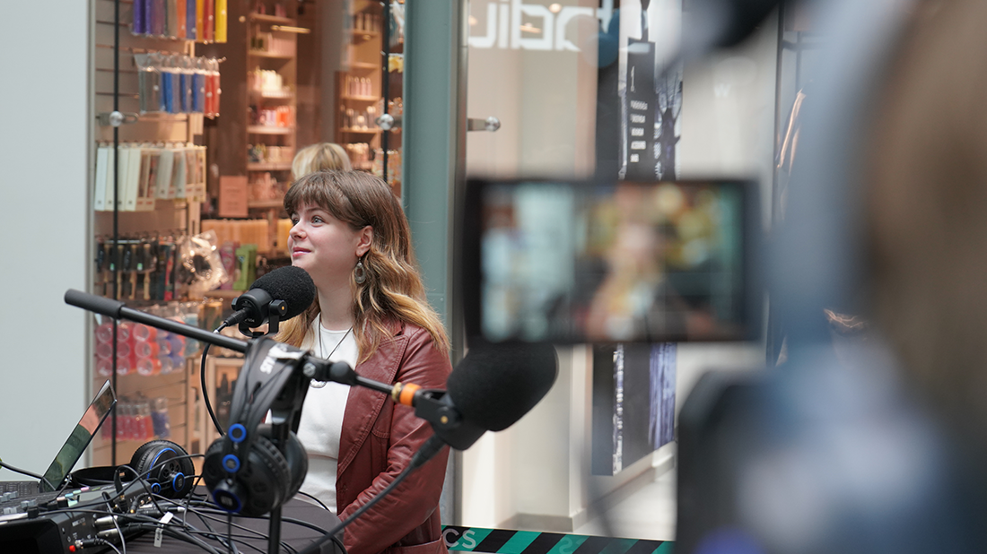 En kvinna står vid en mikrofon, iklädd en vinröd skinjacka och tittar åt sidan och ler.